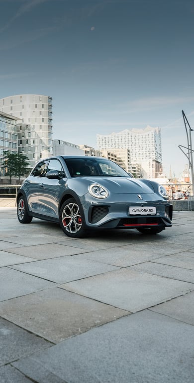 Der GWM ORA 03 GT in Meteorite Grey mit sportlichen Stoßfängern und Leichtmetallfelgen im GT Stil vor der Kulisse der Hamburger Elbphilharmonie