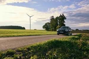 Ein schwarzer GWM ORA 03 fährt bei Sonnenschein über eine schmale Landstraße, rechts von Feldern und Bäumen gesäumt, im Hintergrund steht eine große Windkraftanlage auf einer grünen Wiese.