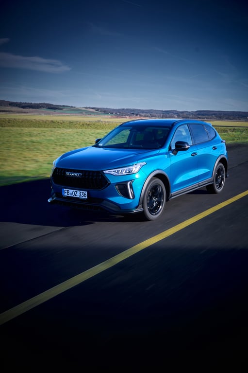 Blauer GWM Haval Jolion Pro SUV fährt auf einer Landstraße durch eine grüne, leicht hügelige Landschaft unter klarem blauem Himmel.